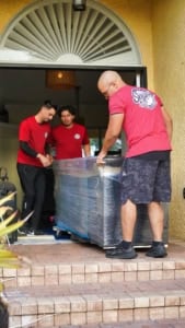 Three movers guide a large, wrapped furniture item out of a home and down the front steps.
