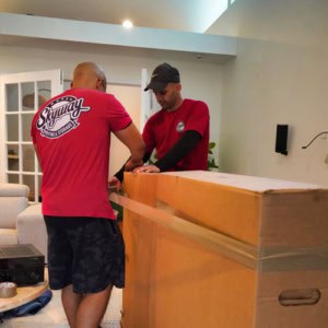 Two movers from Skyway Moving & Storage tape a large cardboard box inside a customer’s living room.