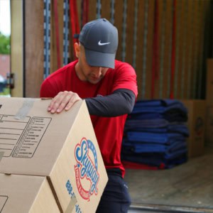 A Skyway Moving & Storage team member in a red uniform and cap lifts a labeled cardboard box into a moving truck.