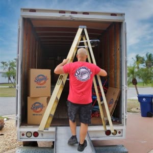 A mover carries a folded ladder up a ramp into a Skyway Moving & Storage truck parked in a residential driveway.
